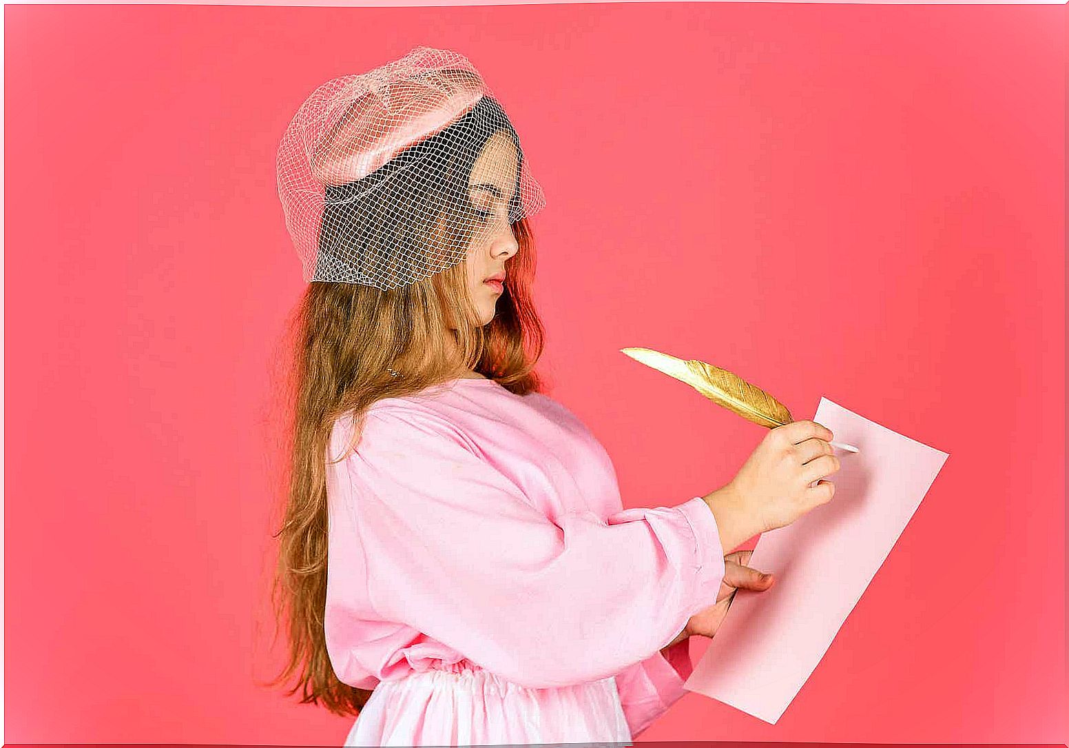 A girl dressed in an old-fashioned hat and dress, writing on a paper with a feather.