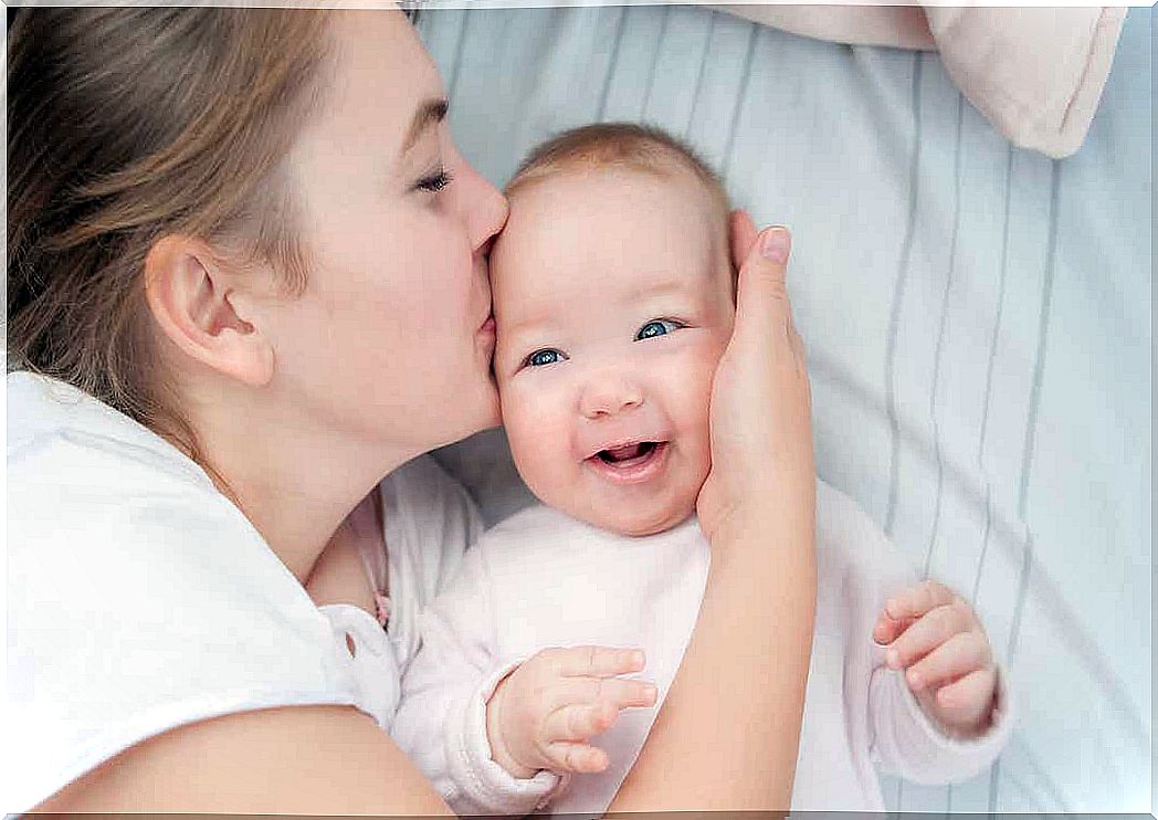 A mother lies in bed with her baby and kisses her on the side of her head.