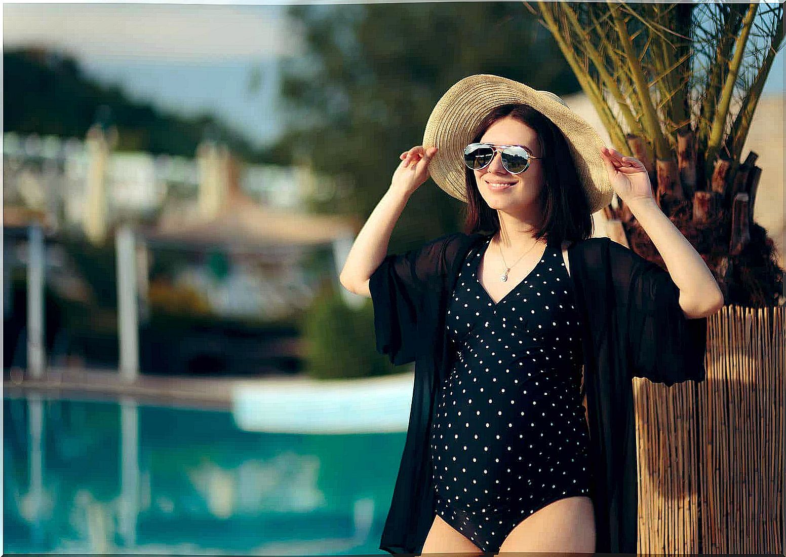 A pregnant woman standing in the sun by a swimming pool.